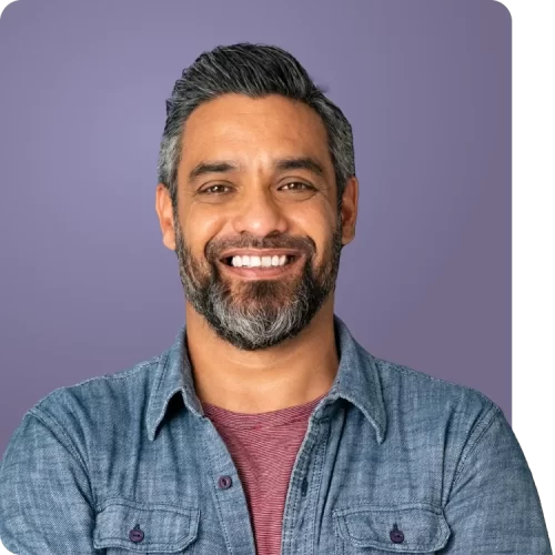 Hombre expresando confianza y bienestar Hombre sonriente con barba y camisa de mezclilla frente a un fondo morado